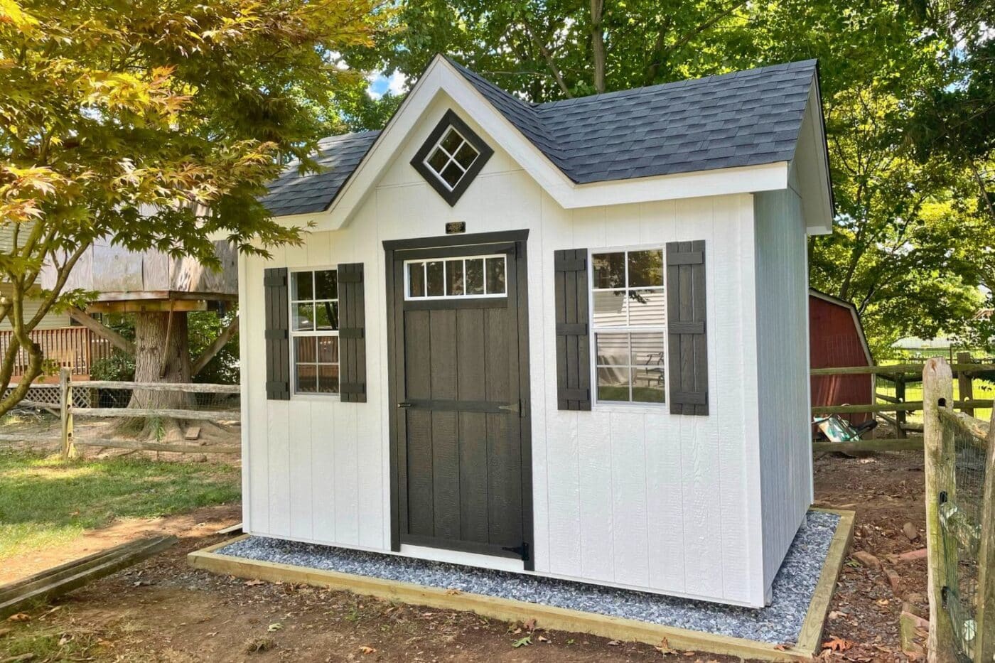 The Cape Cod Shed 2 Wood cape code shed with black doors and shutters on gravel pad in backyard