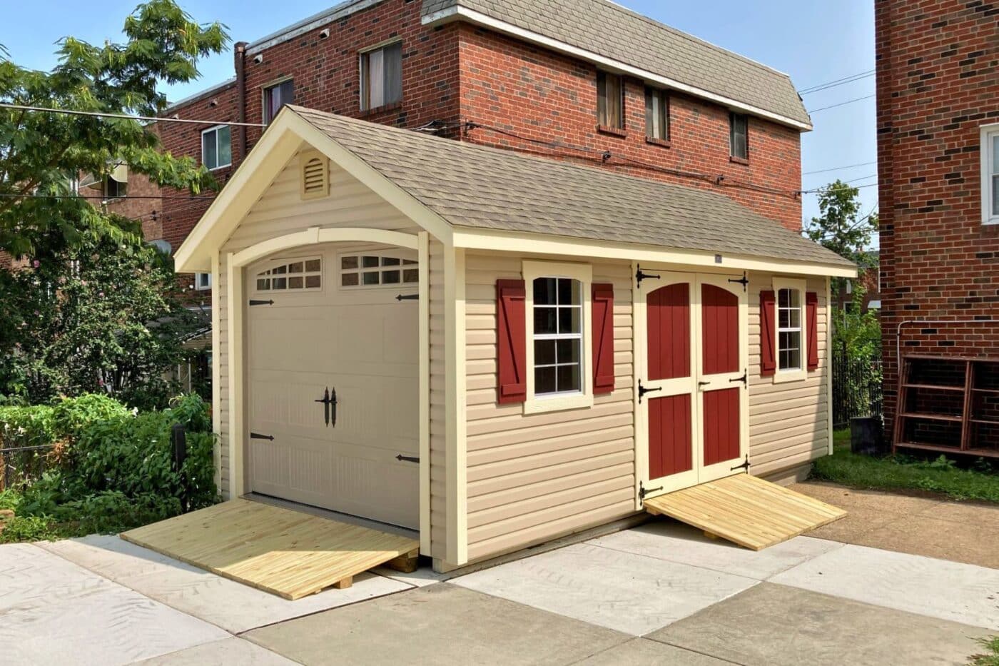 Beige designer workshop garage on concrete patio with ramps and red double doors and window shutters