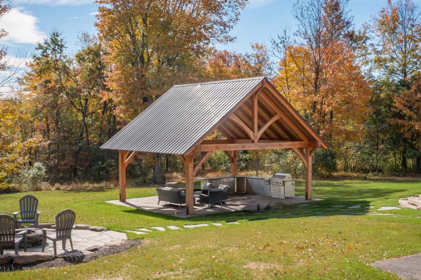 The Kingston Pro TImber Frame Pavilion with seating area underneath. Pavilion is before fal trees and to the right of patio