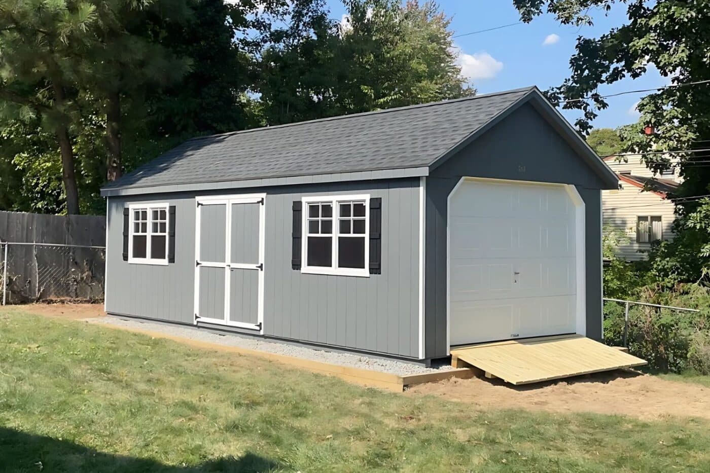 The Standard Workshop Garage 2 Gray garage shed with white trim and doors and black shutters. Shed is set on gravel pad with wooden ramp